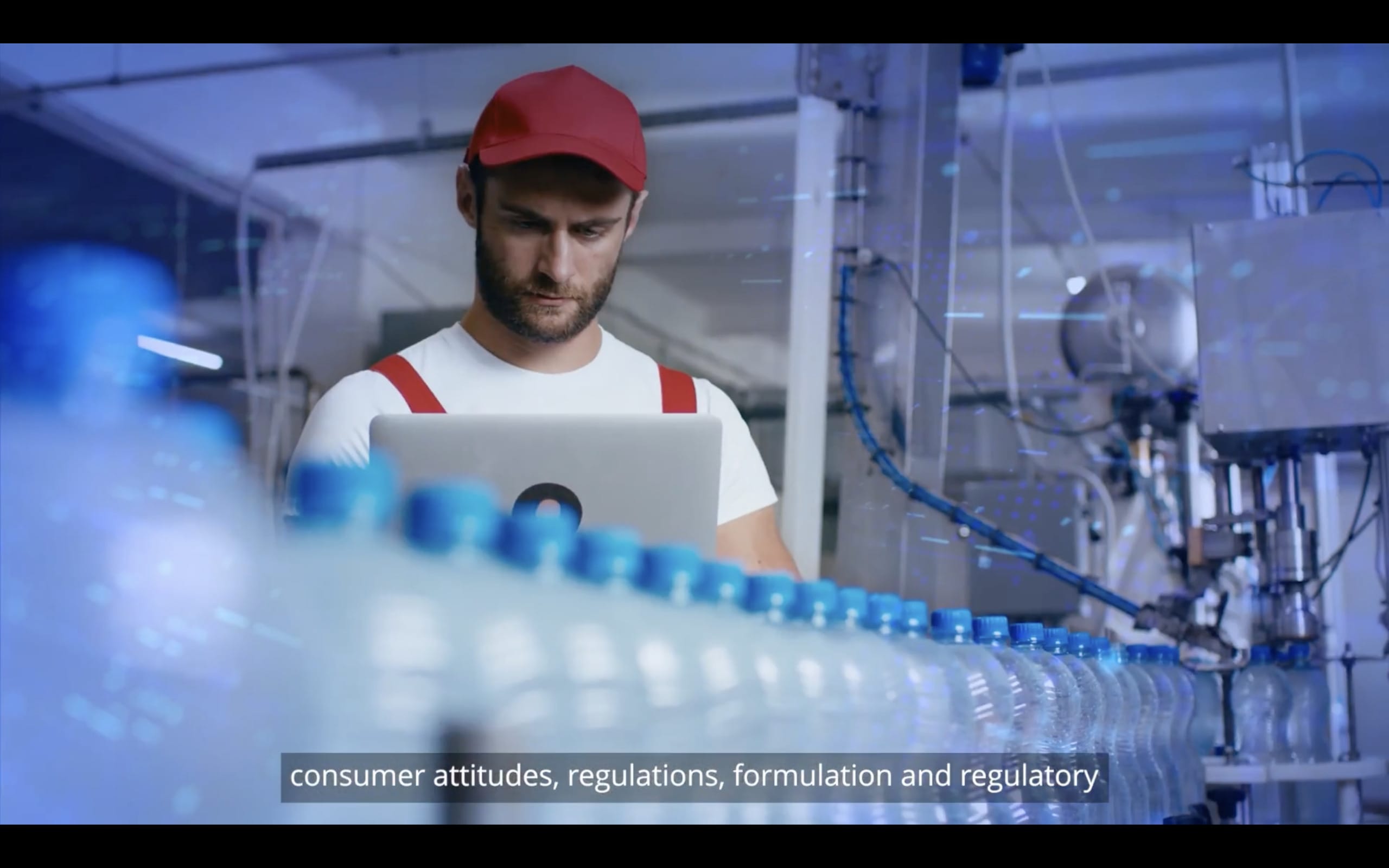 A man in a red cap and white shirt works on a laptop in a bottling factory. Surrounded by machinery, he diligently addresses consumer attitudes and regulations for Complex Beverages. Water bottles, crystal clear, line up in the foreground as subtitles underscore his focus on innovation.