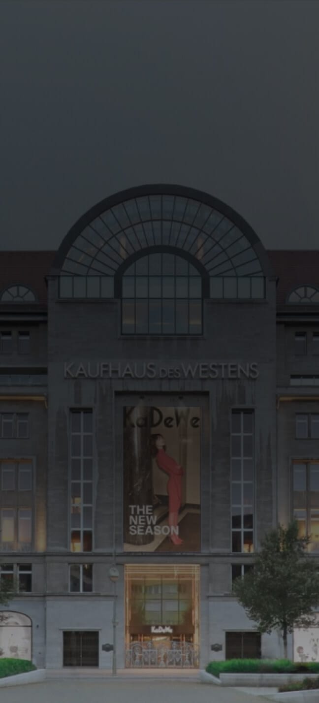 The image captures the entrance of a large department store, "KAUFHAUS DES WESTENS," illuminated at night. A banner with "THE NEW SEASON" fashion ad highlights their centric planning approach. Trees flank the building, adding a touch of nature to this urban spectacle.